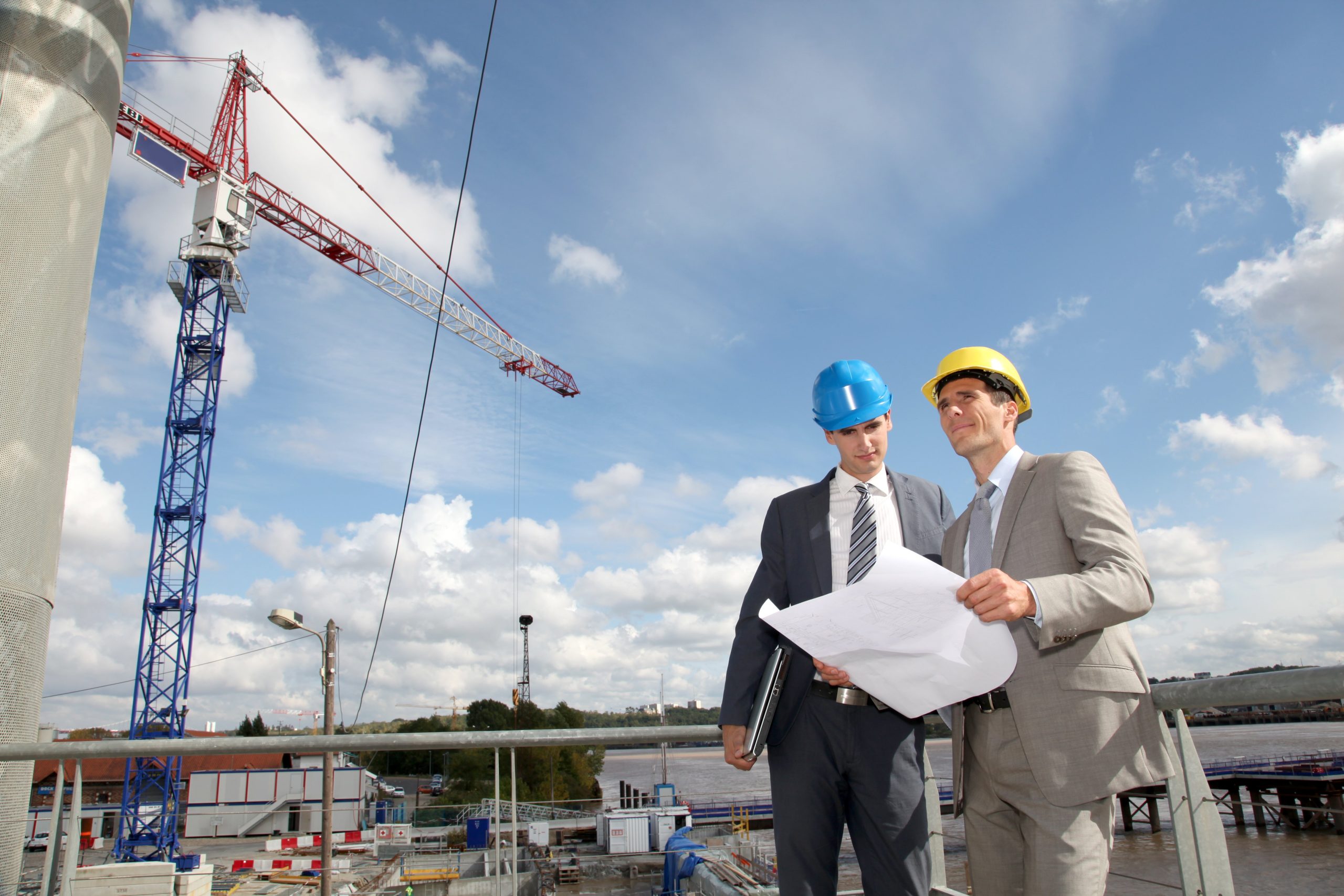 Two professionals reviewing construction plans on-site with cranes in the background.
